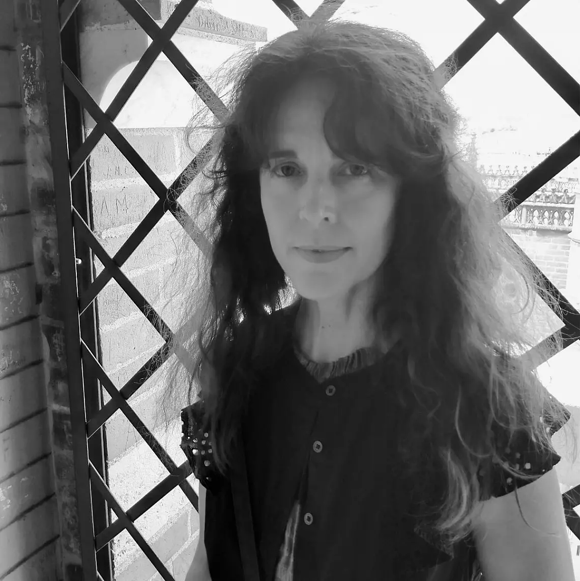 A person with long, wavy hair stands indoors near a window with a lattice pattern. They are wearing a button-up top with studded shoulders. The lighting casts soft shadows, creating a contemplative atmosphere. The image is in black and white.