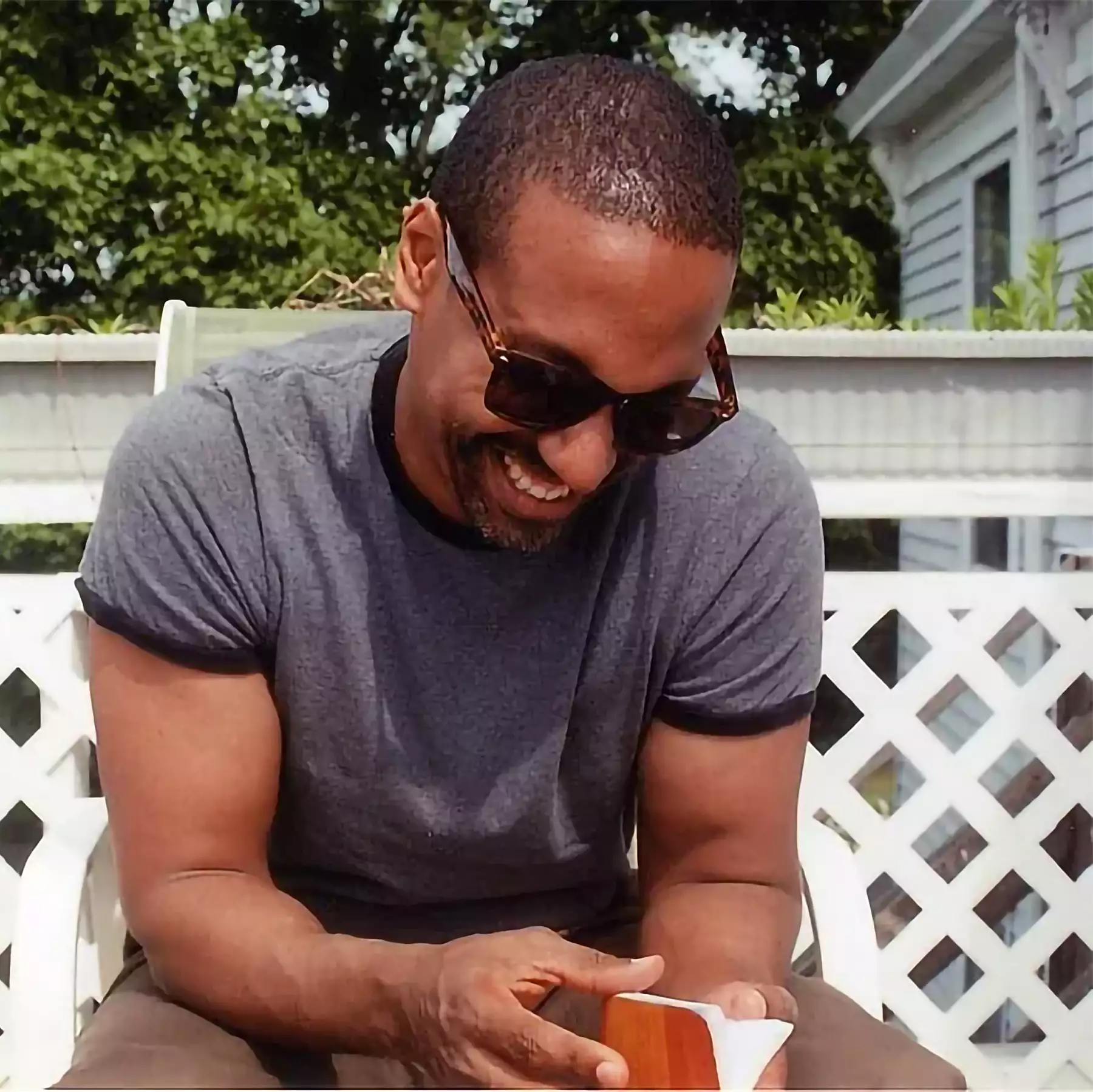Man in sunglasses and a gray t-shirt smiles while sitting outdoors, holding a small brown object. There is a white lattice fence and greenery in the background.