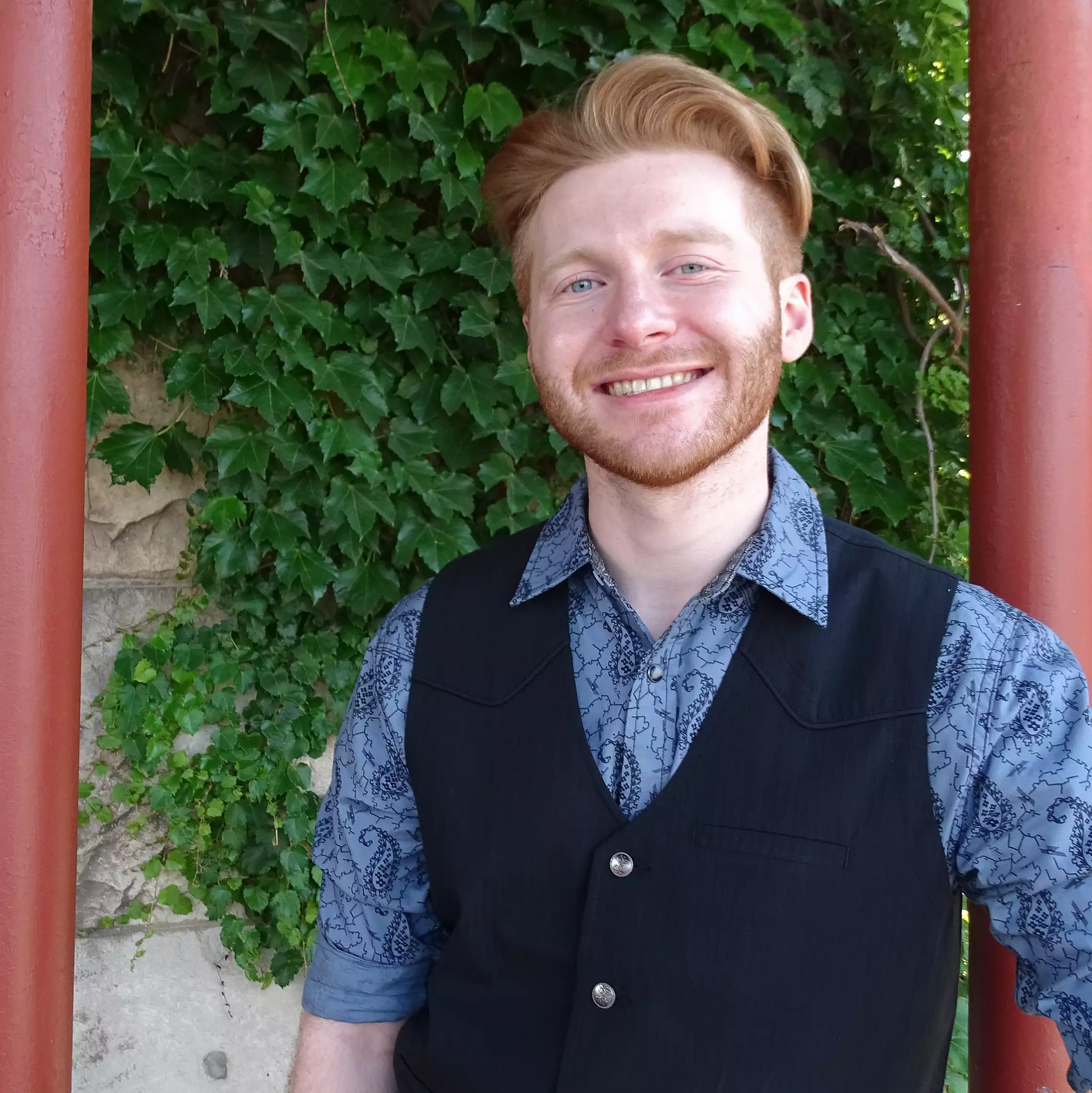 A smiling person with red hair stands outside, framed by red columns. They are wearing a patterned blue shirt and a black vest. Green ivy covers the wall in the background.