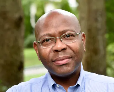 A person wearing glasses and a blue shirt is looking at the camera. They are outdoors with blurred green trees in the background.