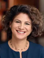 Smiling person with curly hair wearing a blue shirt and necklace.