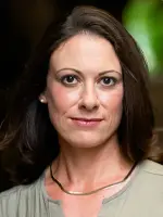 A woman with shoulder-length dark hair and wearing a light-colored blouse and a thin necklace is looking directly at the camera. The background is softly blurred with hints of green.
