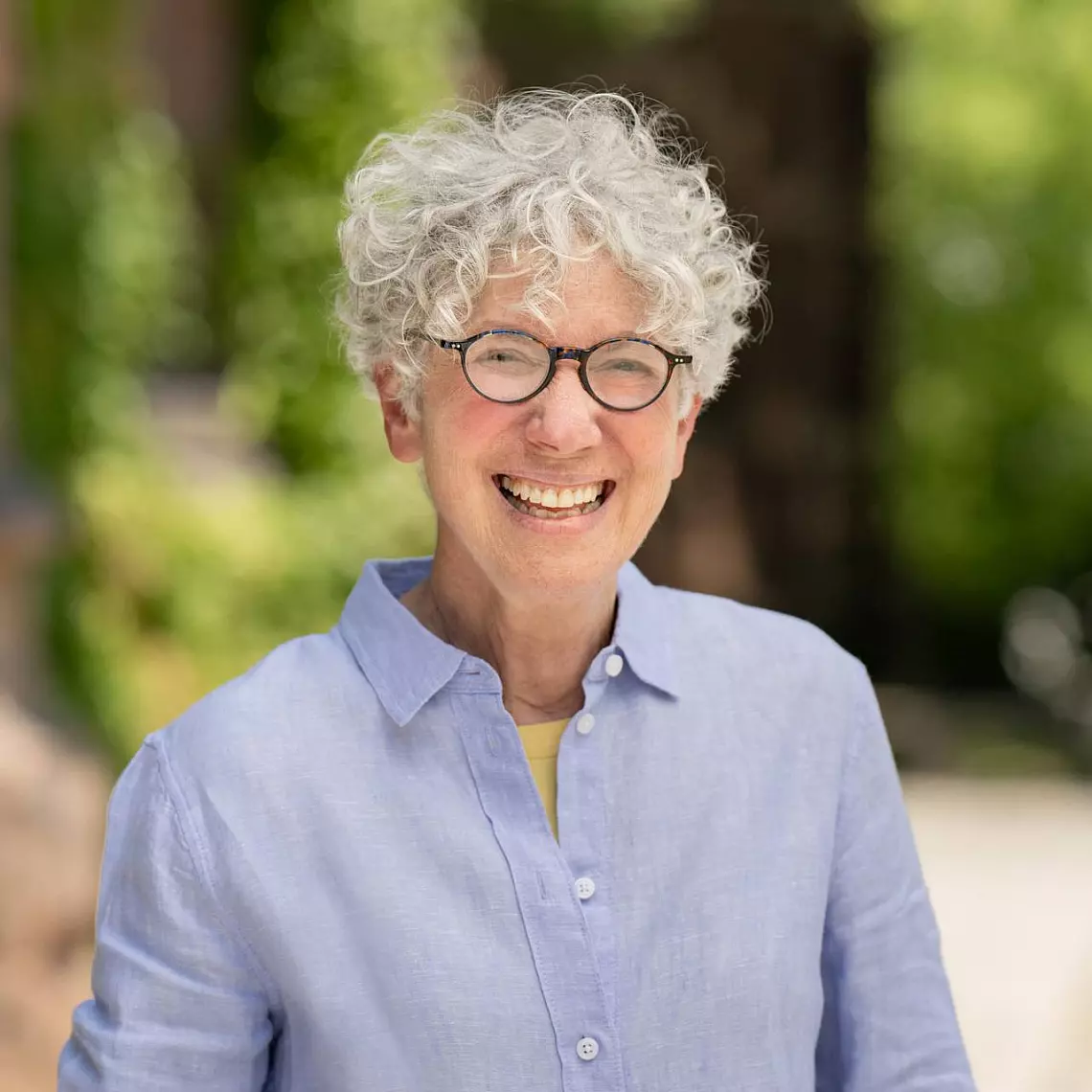 Person with curly gray hair and glasses smiles, wearing a blue button-up shirt. The background is blurry with greenery, suggesting an outdoor setting.