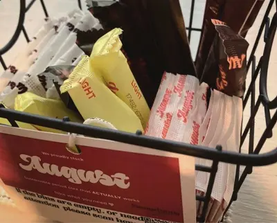 A metal basket containing various tampons and pads in individual colorful wrappers, with a sign that reads "August.