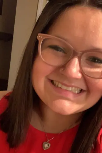 A person with long brown hair is smiling, wearing glasses and a red sleeveless top. They have a pendant necklace and showing bunny ears with their hand. The background shows a partially open door.