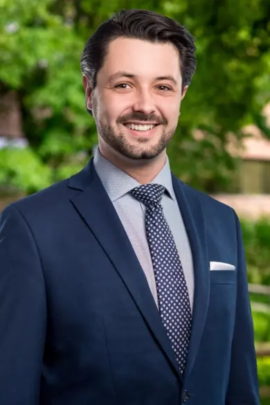 A man with dark hair and a beard is smiling while wearing a navy suit, a light shirt, and a polka dot tie. He is standing outdoors with trees and a building in the background.