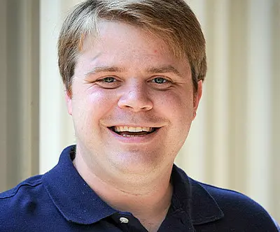 A man with short, light brown hair smiles at the camera. He is wearing a navy blue polo shirt. The background is softly blurred, featuring vertical light-colored lines.