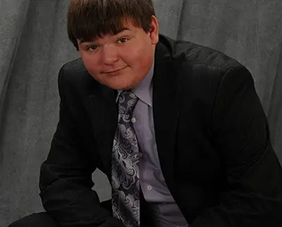 A person in a dark suit, light patterned tie, and lavender shirt sits slightly leaning forward against a gray backdrop, smiling subtly.