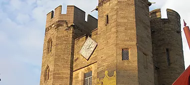 A portion of a historic stone castle featuring two round towers with battlements. Between the towers is a clock face on the stone wall. The sky is clear with a few clouds.