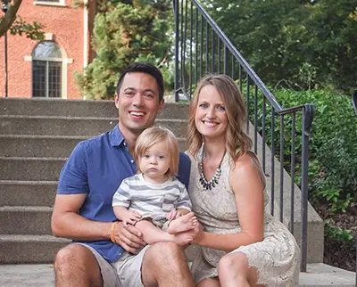 A man and woman sit on stone steps outside, smiling at the camera, with a young child in their lap. They are surrounded by greenery and in front of a red brick building.
