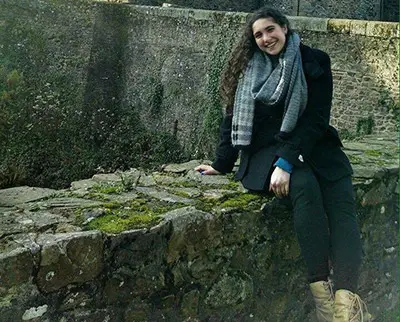 A person with long curly hair, wearing a scarf and coat, sits on a moss-covered stone wall in an outdoor setting. The background features a large stone structure.