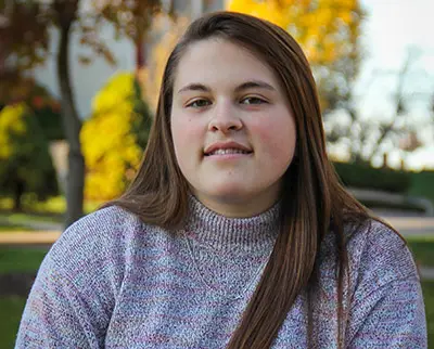 A person with long brown hair wearing a gray sweater is sitting outdoors. The background features trees with autumn leaves and a building.