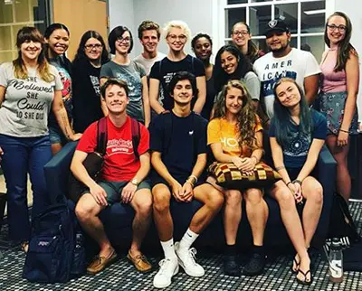 A group of thirteen young adults smiling and posing together indoors. They are arranged in two rows, with some seated on a couch and others standing behind. The setting appears to be a casual, social environment.