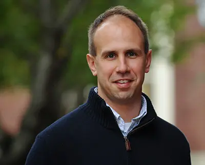 A man with short hair, wearing a dark sweater over a collared shirt, smiles at the camera. He stands outdoors with blurred trees and a building in the background.