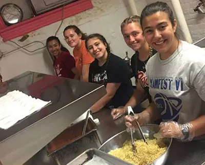Five people smiling while preparing food in a kitchen. They are standing in line, wearing casual clothes, and using gloves to handle ingredients in a large metal bowl. The kitchen has stainless steel surfaces and some kitchenware.