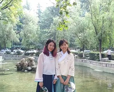 Two women in traditional attire stand smiling by a pond in a park. They hold fans and are surrounded by lush greenery and trees. Cars can be seen in the background.