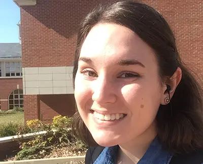 A person with short dark hair is smiling outdoors, wearing a denim shirt and earphones. They are in front of a red brick building under a clear blue sky.