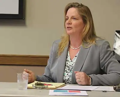 A woman with long blonde hair sits at a table wearing a gray blazer and patterned blouse. She appears to be speaking, with a glass of water, papers, and a clipboard in front of her on the desk.