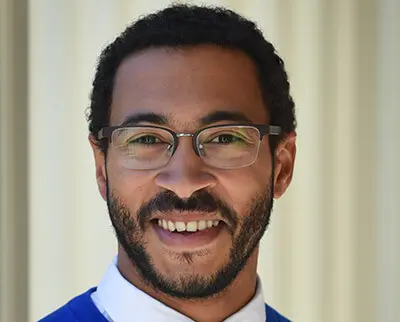 A person with short curly hair and a beard smiles at the camera, wearing glasses, a blue sweater, and a white collared shirt. The background is a light, blurred columned structure.