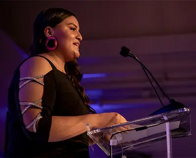 A person with long dark hair speaks at a podium, wearing a black dress with decorated sleeves and large pink earrings, under purple lighting.