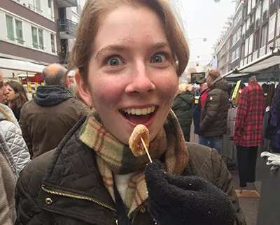 A person with a surprised expression is being fed a treat on a skewer in an outdoor market. They're wearing a checkered scarf and a dark jacket, with people and market stalls in the background.