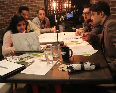 A group of people sit around a table in a dimly lit room, engaged in discussion. Laptops, papers, and drinks are scattered across the table. A brick wall and a modern light fixture are in the background.