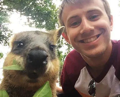 A smiling person takes a selfie with a quokka, a small marsupial, as it eats a leaf. The photo is taken outdoors, with green foliage visible in the background.
