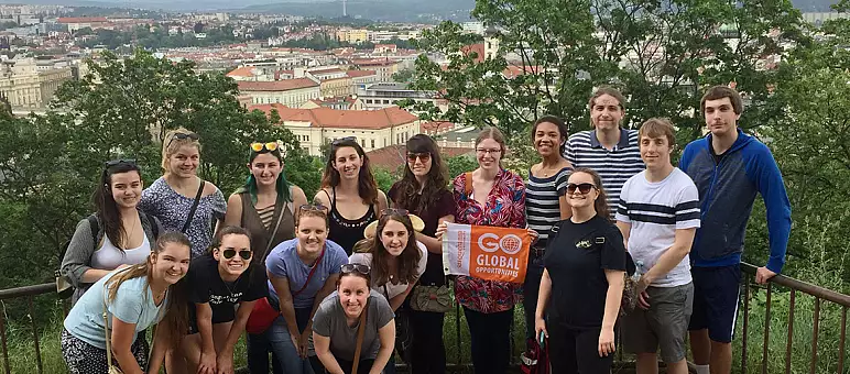 A group of people posing on a scenic overlook with a panoramic view of a city below. They are holding a "Go Global" banner. The city features red-roofed buildings and surrounded by greenery. The sky is cloudy.