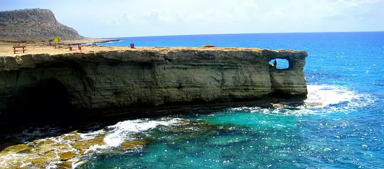 Rocky coast with a natural arch over turquoise waters, beneath a clear blue sky. Sparse vegetation tops the cliff, with picnic tables visible. Waves gently crash against the rock formations below.