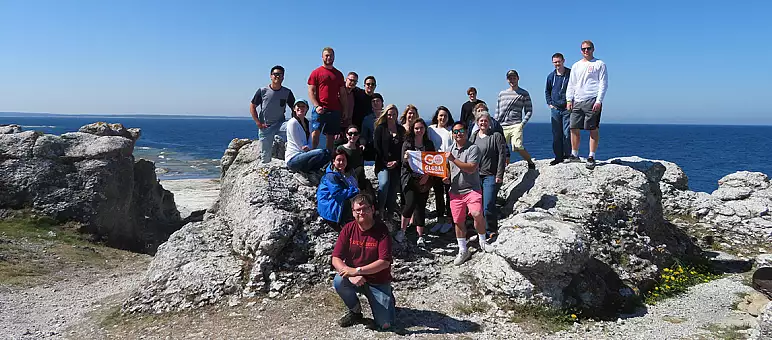 A group of people posing on large rocks with the sea in the background. The sky is clear and blue. One person holds an orange sign that reads "GO". They all appear casually dressed, enjoying a sunny day by the ocean.