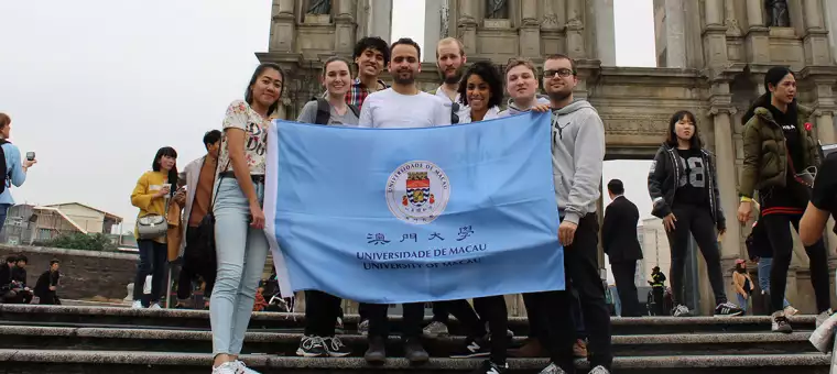 A group of people stand on steps holding a University of Macau flag. They are in front of the Ruins of St. Paul's in Macau. The scene is lively, with several other people in the background.