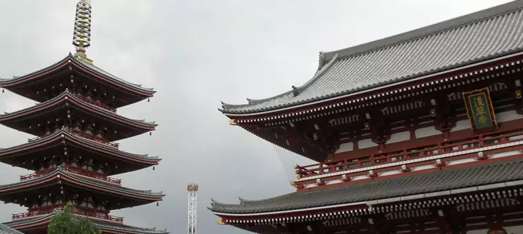A traditional Japanese temple scene featuring a multi-tiered pagoda on the left and an ornate temple building on the right, both with gray-tiled roofs and intricate wooden details, set against a cloudy sky.