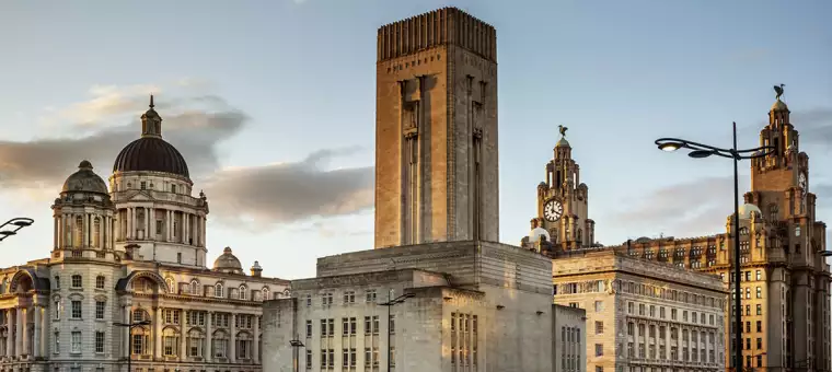 View of Liverpool's iconic skyline featuring historic buildings at sunset. The Royal Liver Building with its clock tower is prominent, alongside other architectural landmarks, under a sky with scattered clouds.
