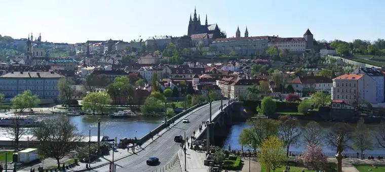 A panoramic view of a city with a bridge over a river in the foreground. The cityscape features dense clusters of buildings and a large castle on a hill in the background. The scene is set on a clear day with trees and greenery.