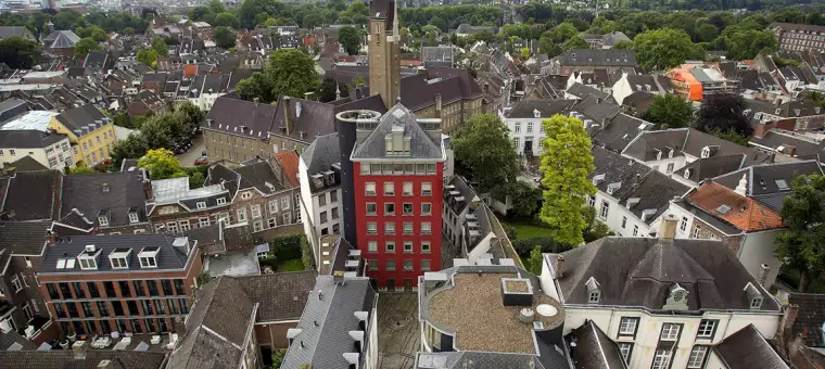 Aerial view of a European cityscape with a mix of old and modern buildings. A prominent red building stands in the center. The surrounding architecture includes trees and various roof styles, with lush greenery in the background.