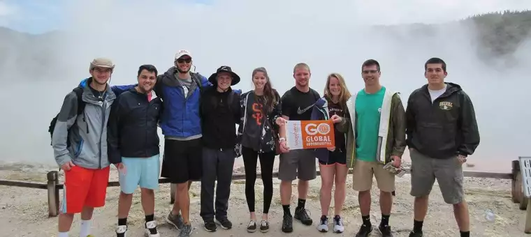 A group of nine people stand together outdoors, smiling at the camera. They hold a sign that says "Go Global." The background features a misty, geothermal landscape.