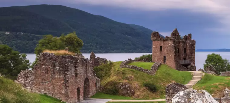 The image shows the ruins of Urquhart Castle, with its tall, partially preserved tower and stone walls, surrounded by lush greenery. The castle overlooks the calm waters of Loch Ness, with a forested hill in the background under a cloudy sky.