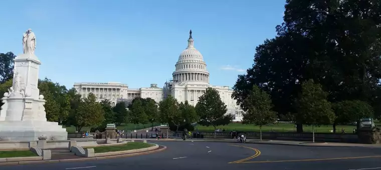 View of the U.S. Capitol building with its iconic dome under a blue sky. Statues and lush green trees frame the scene. The foreground features a curved road and people strolling in the area.