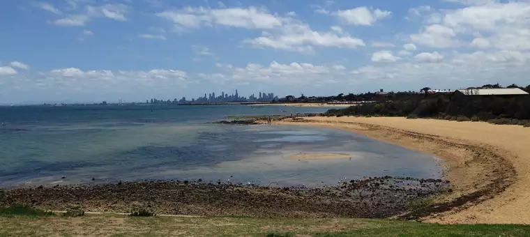 Coastal view with a calm bay and a sandy beach in the foreground. The city skyline is visible in the distance under a partly cloudy sky. Small waves gently lap against the shore, and the landscape is dotted with low vegetation.