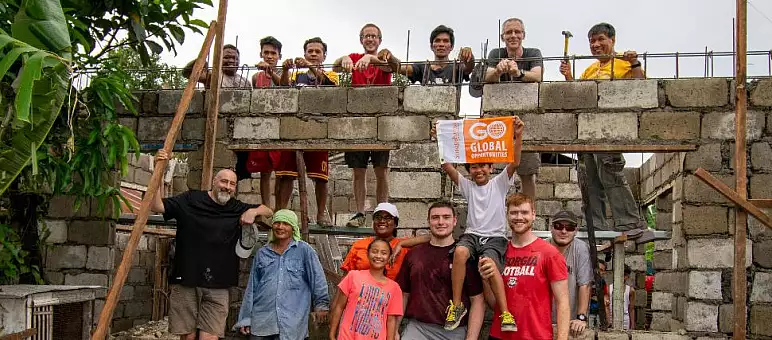 A diverse group of people stand on and around a partially-built brick structure. Some hold tools, and one person holds a sign that reads "Go Global Institute." They appear cheerful and are surrounded by greenery under a cloudy sky.