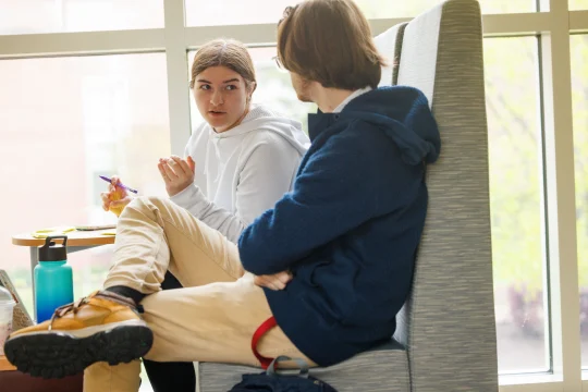 Two people sitting on gray chairs in a brightly lit room with large windows. The person on the left is gesturing while speaking, wearing a white hoodie. The person on the right listens attentively, wearing glasses and a dark blue jacket.