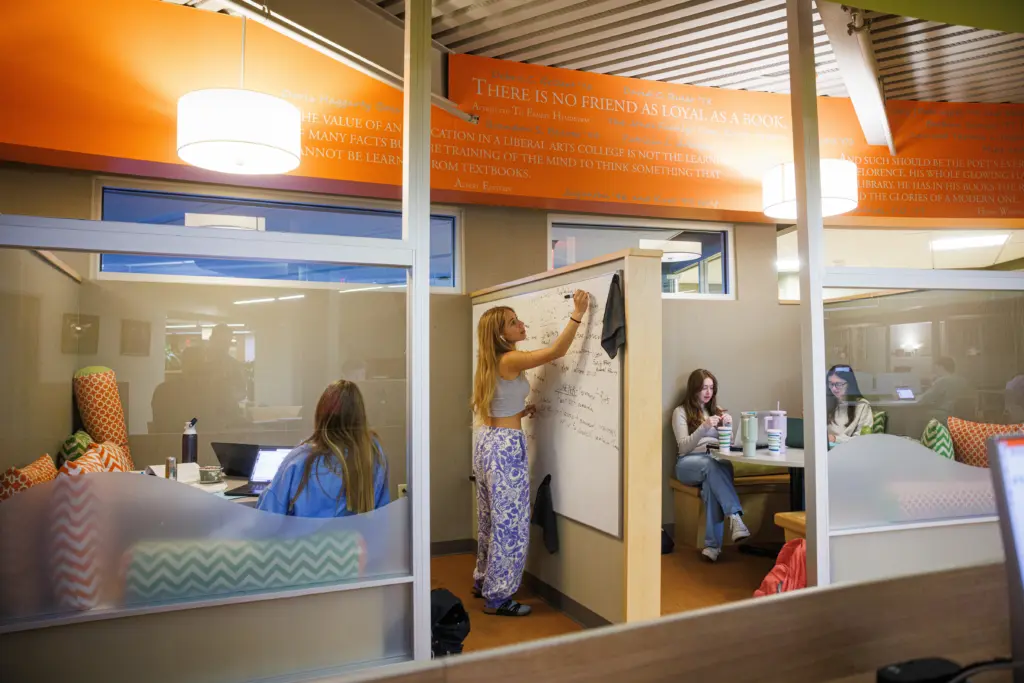 A student writes on a whiteboard inside a glass-walled study area. Other students sit around on couches with laptops and notebooks. The room has orange accents and inspirational quotes on the walls.