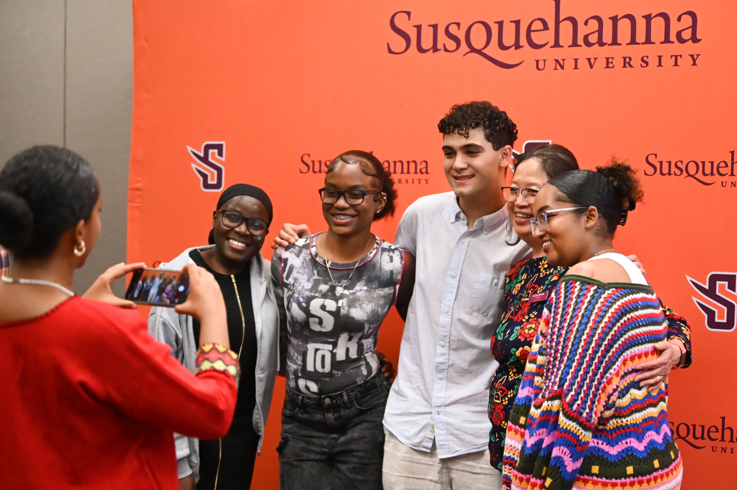 A group of five people are posing and smiling for a photo in front of an orange Susquehanna University backdrop. A person in a red outfit is taking their picture with a smartphone.