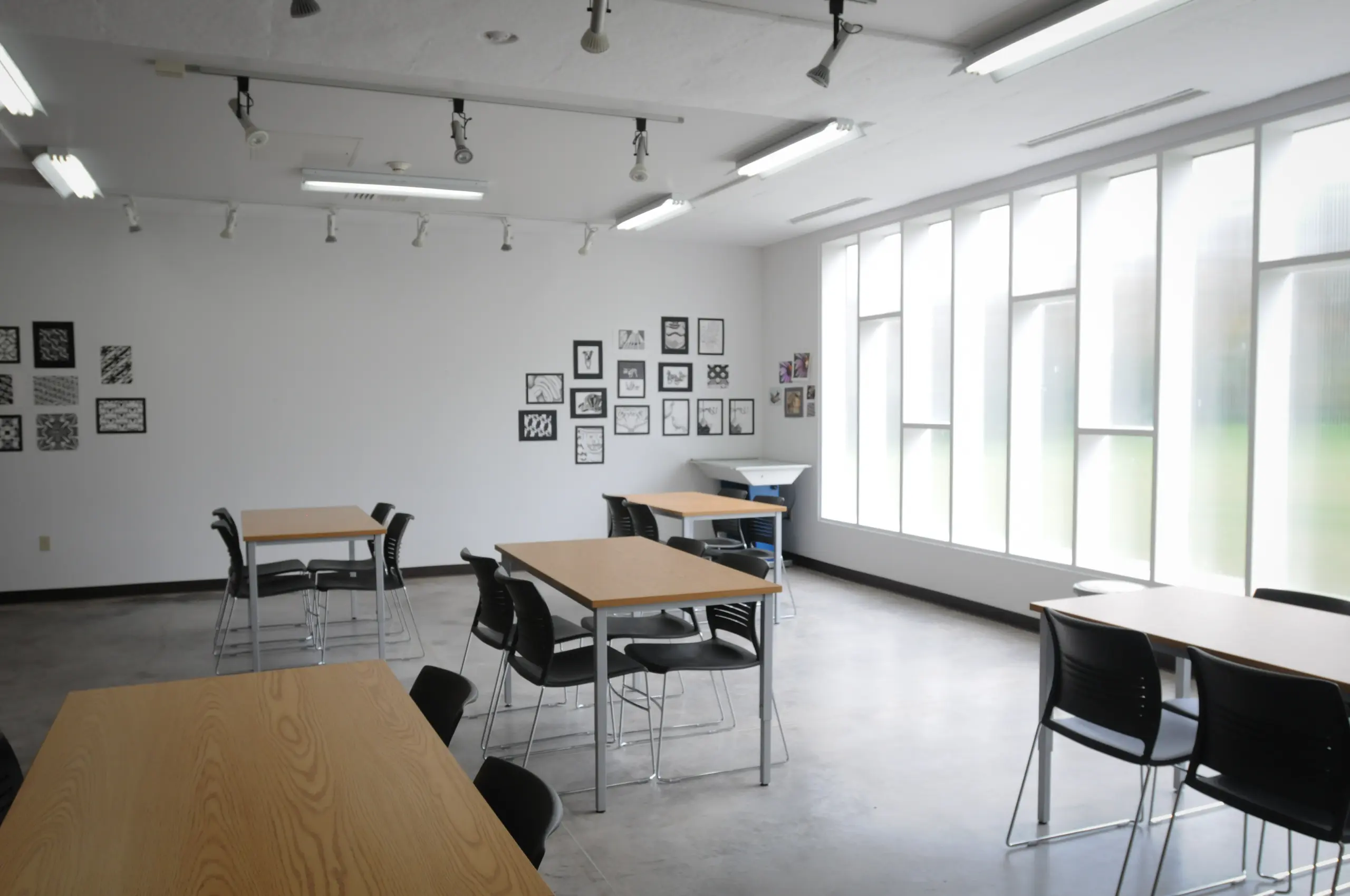 A spacious classroom with large windows allowing natural light. It has wooden tables and black chairs arranged in groups. Framed artwork is displayed on one white wall. The floor is gray and the ceiling has track lighting.