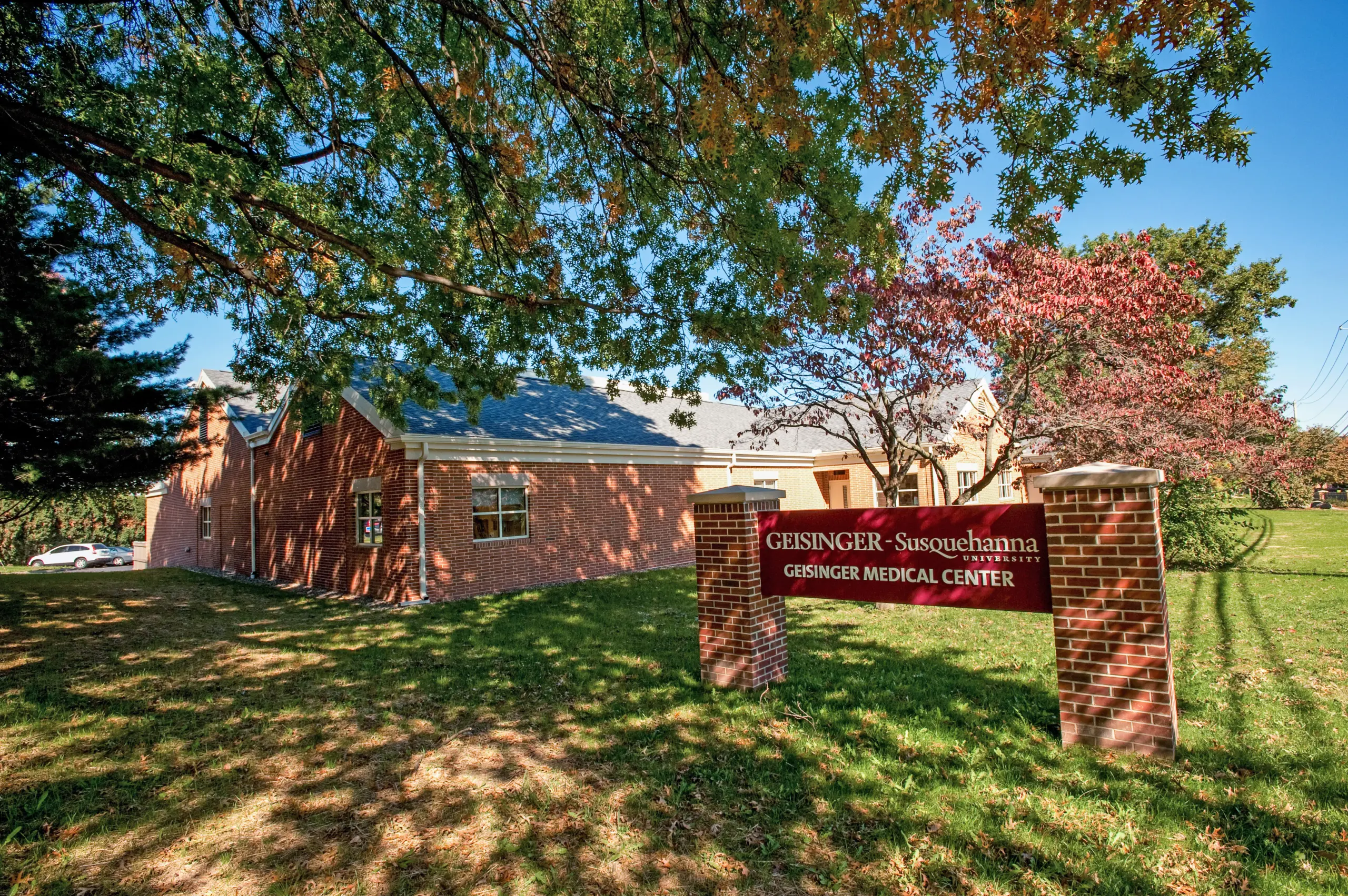 A brick building under a clear sky, surrounded by trees with autumn leaves. A sign in front reads "Geisinger Susquehanna, Geisinger Medical Center." The grass is green, with patches of sunlight filtering through the branches.
