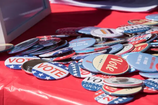 A collection of round, colorful buttons scattered on a red tablecloth. The buttons feature words like "VOTE" and have patriotic designs with stars and stripes, promoting participation in elections.