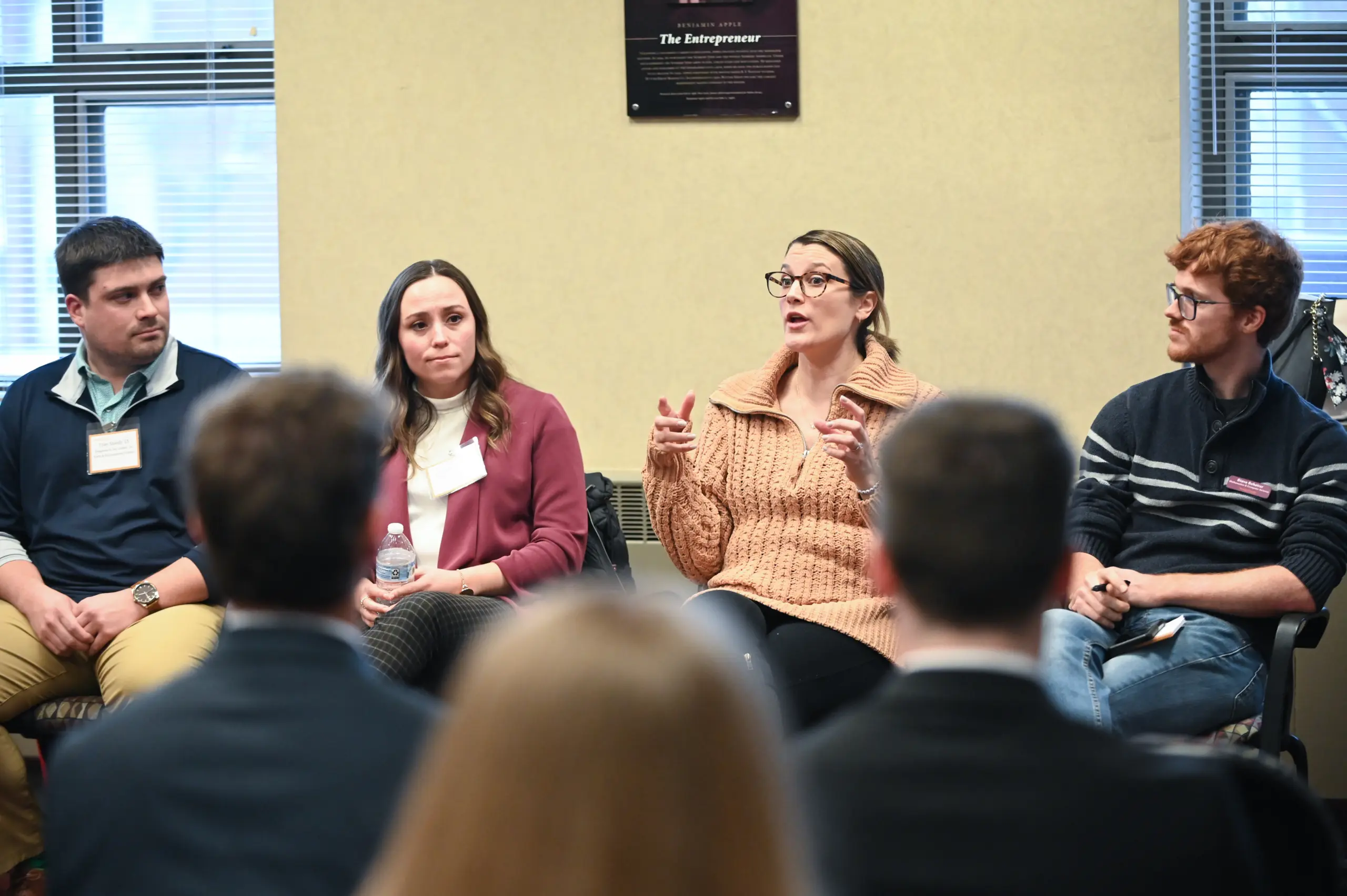 Four individuals sitting in a panel discussion in a classroom setting. A woman in a brown sweater speaks while gesturing with her hands, flanked by two men and a woman. Audience members are visible in the foreground.