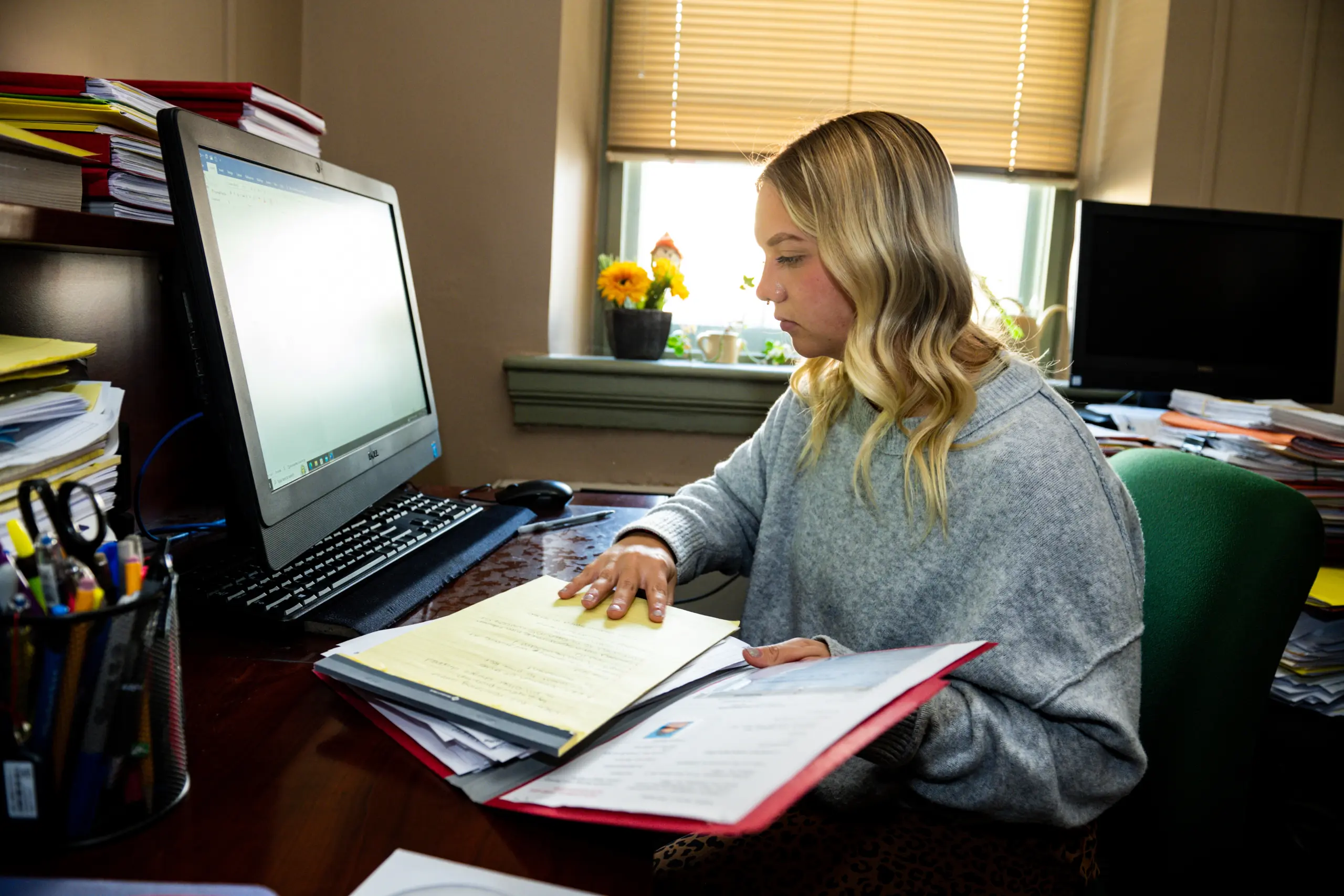 A woman with blonde hair sits at a desk, working on documents in a brightly lit office. A computer monitor and stack of folders are in front of her. The window behind her has blinds and a small plant with yellow flowers on the sill.