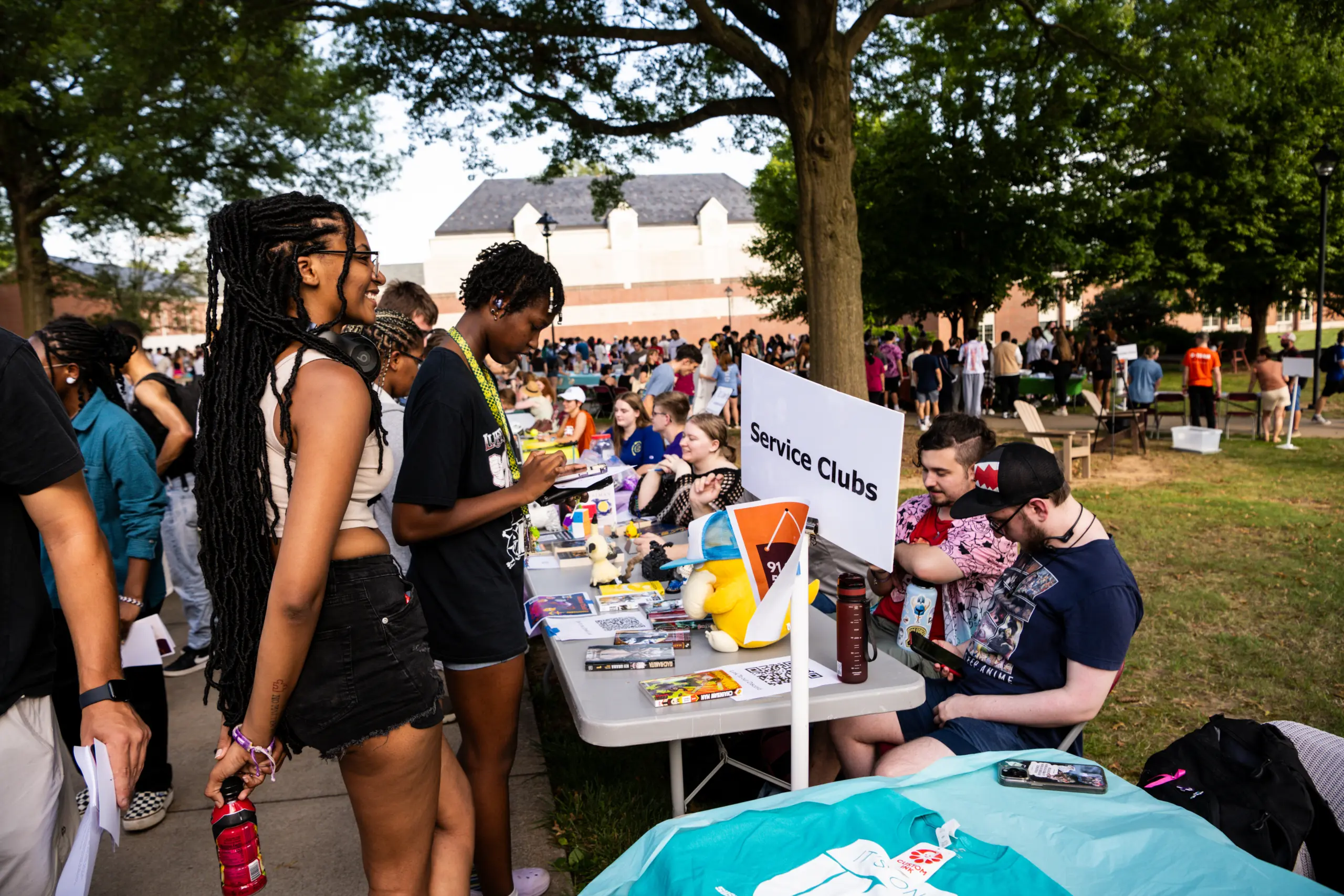 People are gathered outdoors at a community or school event. A table labeled "Service Clubs" is set up with various items on display. Attendees are interacting, and the scene is lively with trees and buildings in the background.