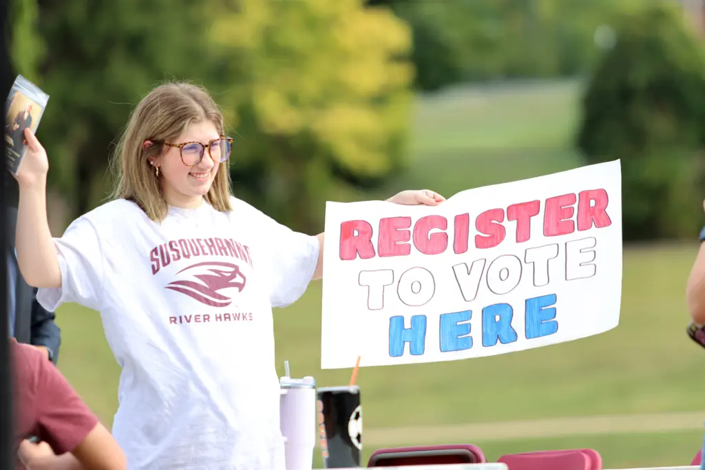 A person wearing glasses and a "Susquehanna River Hawks" shirt holds a sign that reads "Register to Vote Here" in red and blue letters. They are outside, with trees and grass in the background.